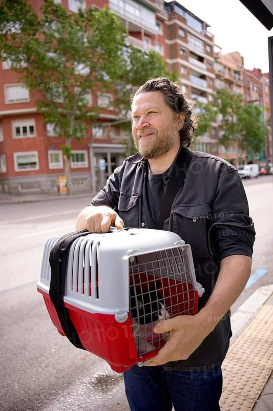 A man carrying a red and white pet carrier with a cat, walking along a city street. The background shows apartment buildings with trees lining the sidewalk