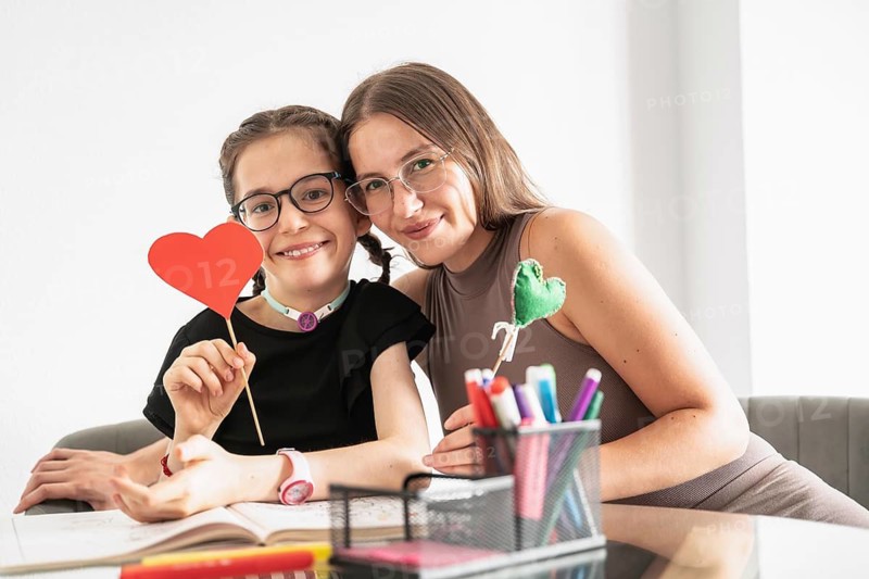 A joyful family moment featuring a young girl with a tracheostomy, surrounded by love and creativity. She holds a heart symbol, highlighting the nurturing bond and inclusive environment