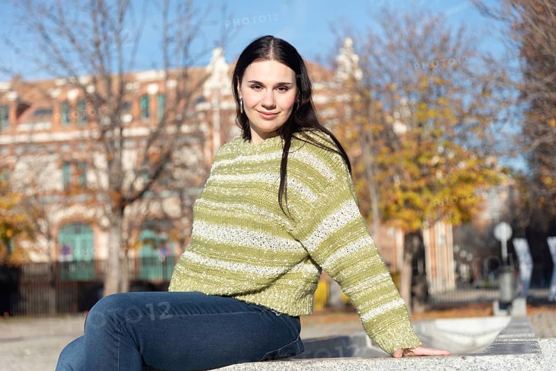 A woman with dark hair sits casually on a concrete bench, wearing a green sweater and jeans. The sunny day highlights the colorful foliage around her