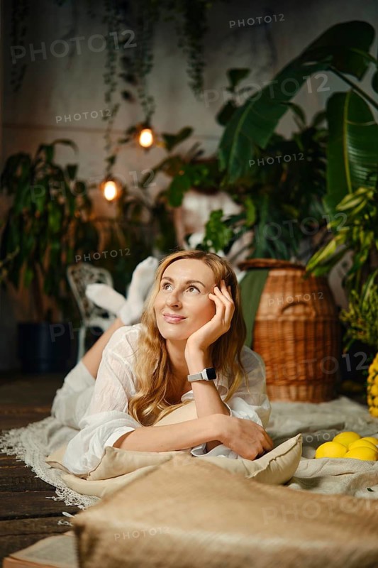 Dreamy woman rests on a soft rug with her chin on her hand, looking up in thought. Green plants surround her, and there are fruits nearby. The light is warm, creating a cozy atmosphere