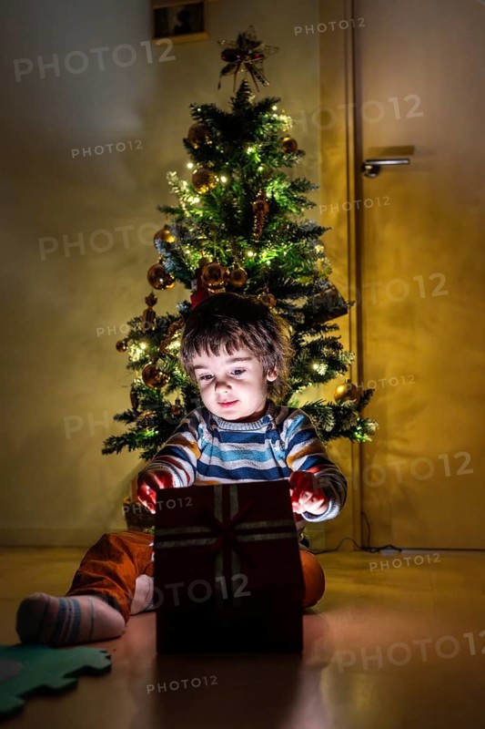 A young child sits in front of a Christmas tree, opening a red present. The scene is warm and festive, with the child's excitement and the tree's decorations creating a joyful atmosphere