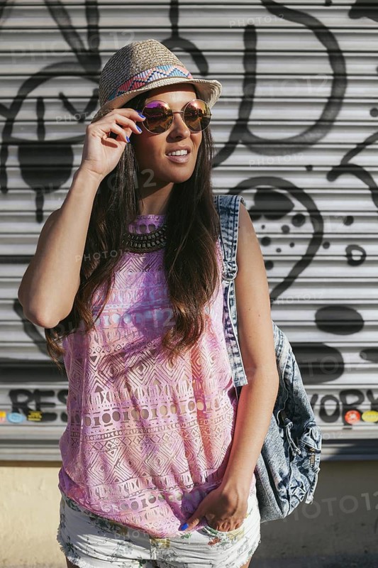 Stylish woman wearing a straw hat and sunglasses stands confidently in front of urban graffiti. Casual summer attire with a denim vest and patterned top