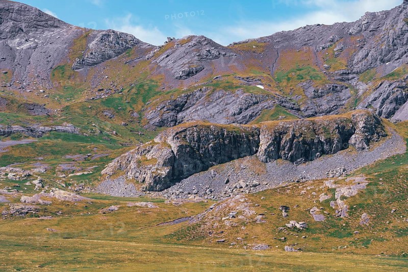 Serene view of the lush and rugged terrain of the Ibones de Anayet located in Huesca, Spain, showcasing vivid green meadows and imposing rocky mountain peaks under a clear blue sky