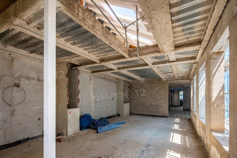 A spacious interior construction site featuring exposed metal framework and unfinished walls. Sunlight filters through large windows, illuminating tools and building materials