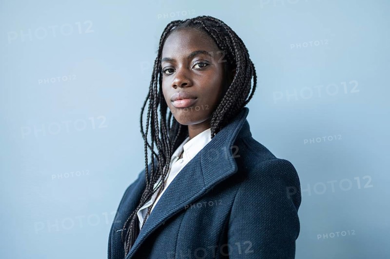 African American businesswoman with braids, dressed in a formal suit, poses against blue background, reflecting professionalism and confidence