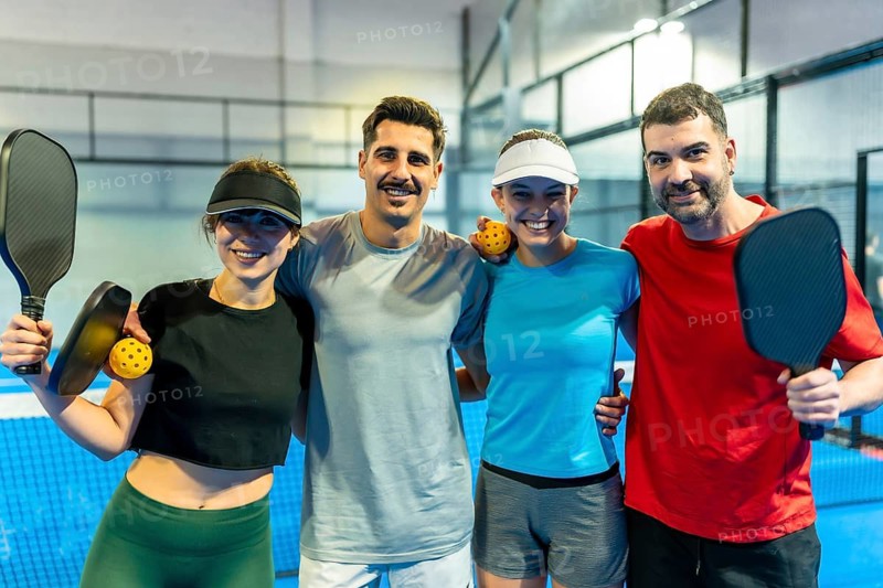 Group of four mixed gender pickleball players embracing and smiling, holding equipment on a blue indoor court after a game, symbolizing teamwork and healthy lifestyle