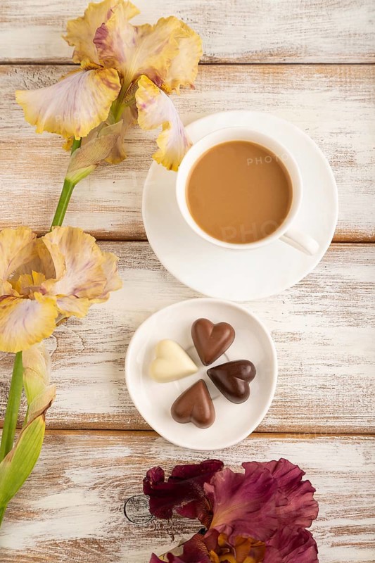 Cup of cioffee with chocolate candies, lilac and purple iris flowers on white wooden background. top view, flat lay, close up, still life. Breakfast, morning, spring concept