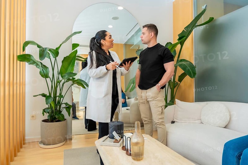 Doctor engaging in a discussion with a male patient, holding a tablet and providing information during a consultation in a contemporary aesthetic center waiting area