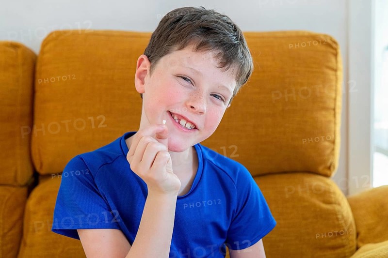 A joyful young boy sits on a sofa, showing off his newly lost milk tooth, with a big smile, indicating a milestone in his childhood