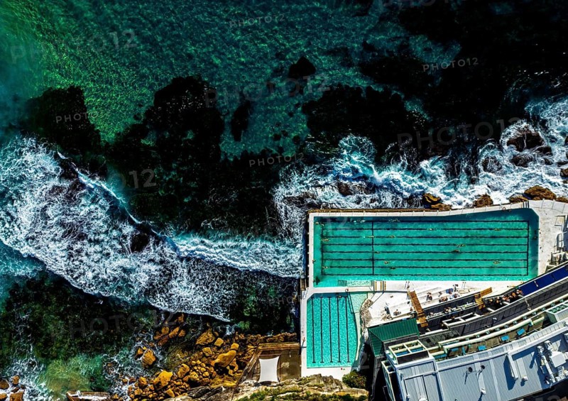 A aerial shot of a swimming pool along a rocky coastline in Sidney, Australia. The turquoise water contrasts with the white waves crashing into the rocks, creating a scenic coastal view