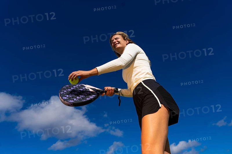 From below a young woman in sportswear is captured in the act of playing tennis against a bright blue sky background, focusing intently on her next shot