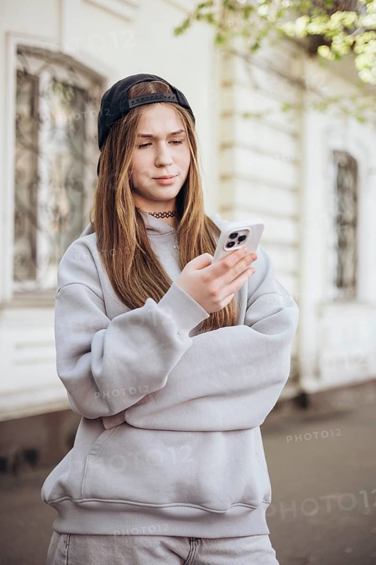 A teenage girl in casual clothing stands outside, focused on her smartphone. She wears a cap and hoodie, indicating a relaxed, modern lifestyle connected by technology