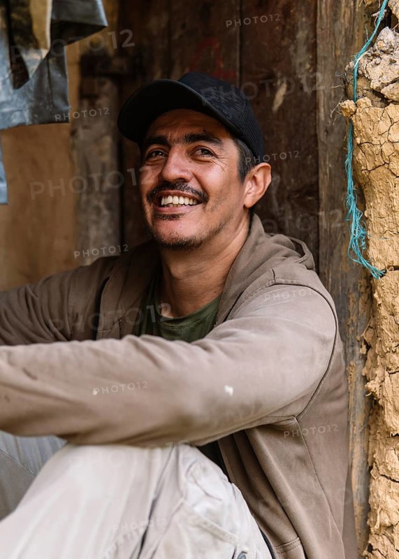 A cheerful man wearing a cap and jacket sits comfortably in a rustic setting in rural Colombia, surrounded by natural textures and earthy tones, exuding joy and simplicity