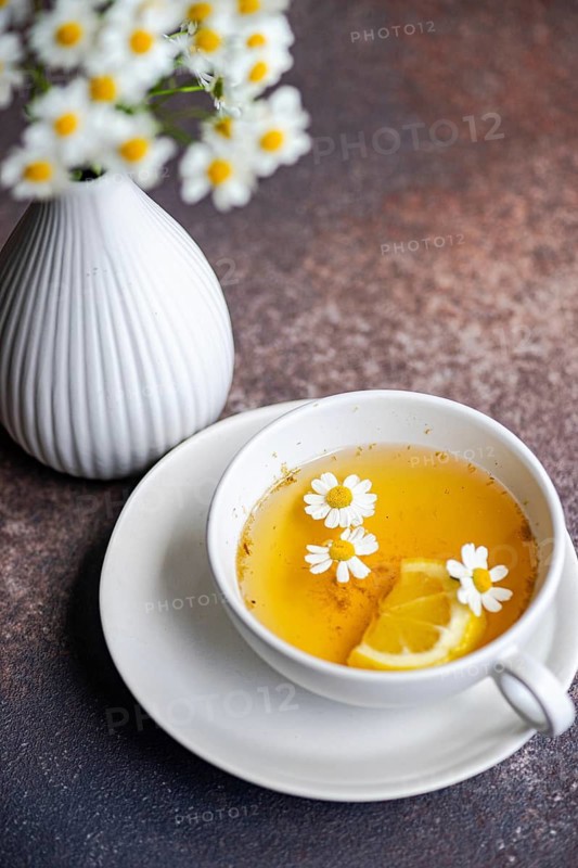 A white vase filled with summer daisies sits beside a cup of chamomile tea garnished with lemon and flowers. The arrangement rests on a textured brown surface