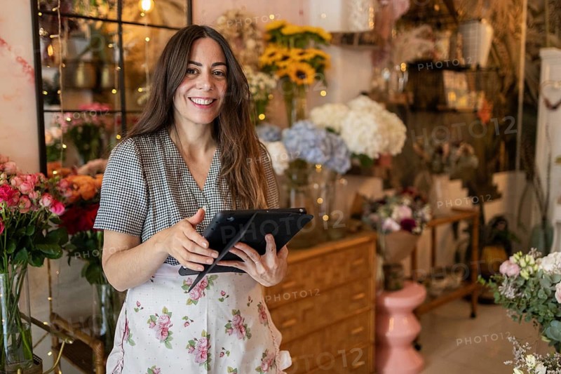 A smiling woman in a cozy flower shop holds a tablet while surrounded by vibrant flowers. The warm ambiance showcases blooming arrangements and rustic decor, creating an inviting atmosphere