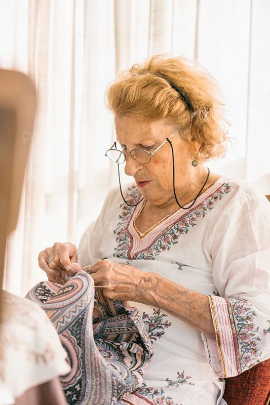 Concentrated senior woman wearing eyeglasses and casual clothes carefully sewing colorful patterned fabric while sitting on chair in cozy room at home