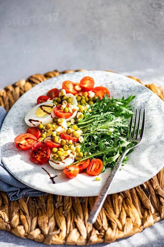 A vibrant keto lunch featuring boiled eggs, fresh rocket, cherry tomatoes, canned green peas, and sprouted green beans, all drizzled with balsamic sauce for added flavor