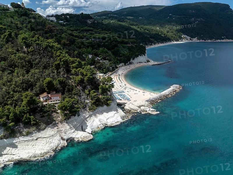Stunning aerial view of Sirolo beach, Italy, showcasing turquoise waters, sandy shorelines, and lush green cliffs under a partially cloudy sky