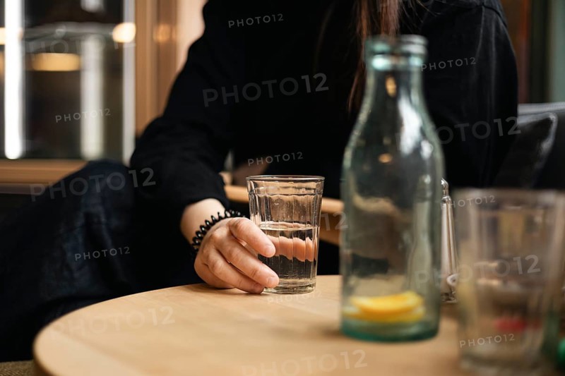 Cropped unrecognizable girl holding a glass of water at a cafe table, with a bottle beside her and a cozy ambiance