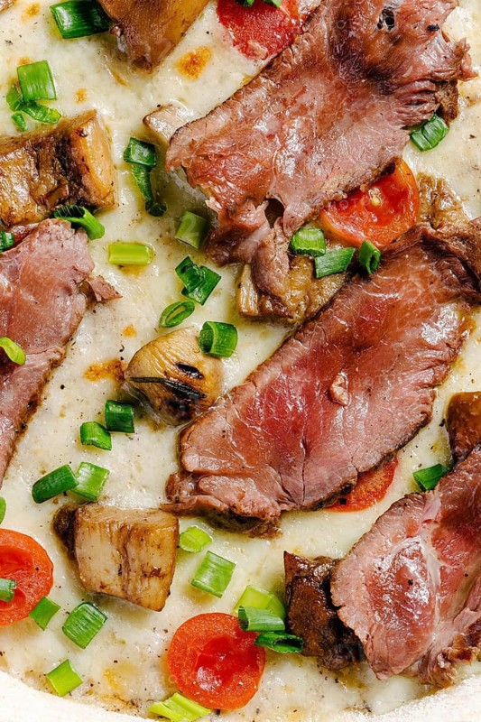 Close-up view of mozzarella pizza topped with roast beef, porcini mushrooms, cherry tomatoes, and sprinkled with green onions