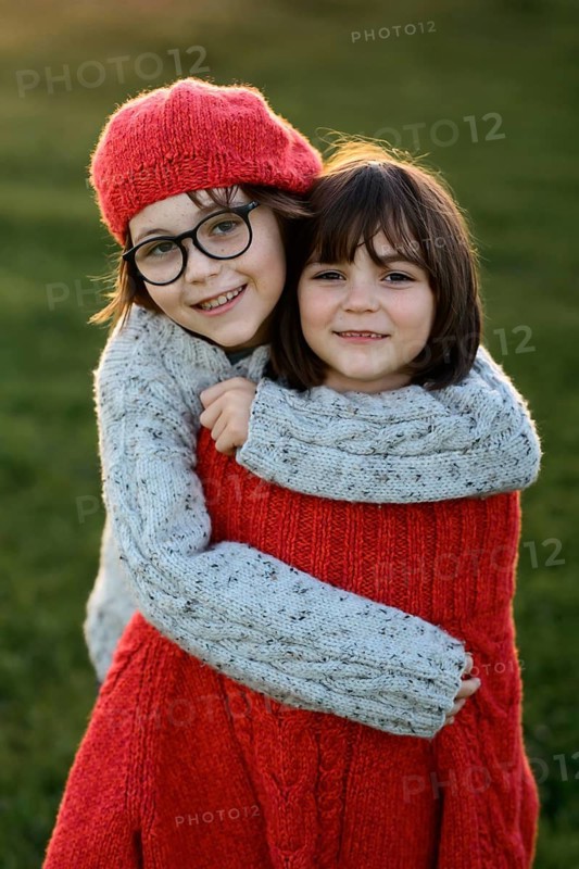 A girl and a boy warmly embrace, dressed in cozy knitted sweaters and caps during a picturesque autumn sunset. Their joyful connection radiates against the soft, natural light