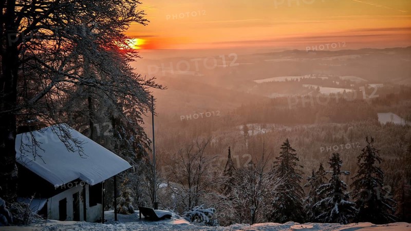 Snowy hut in a wintry mountain landscape at sunset, surrounded by trees and warm light, view from Döbra Berg, wide winter landscape with spectacular orange sunset with snow and trees, Frankenwald nature park Park