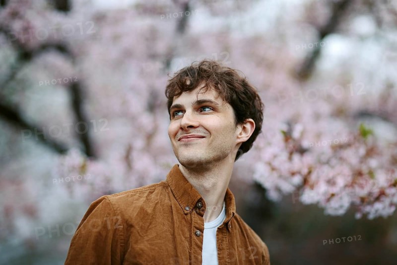 A cheerful man in a brown jacket smiles beneath cherry blossoms in full bloom during springtime in Japan, illustrating the joy and beauty of sakura season