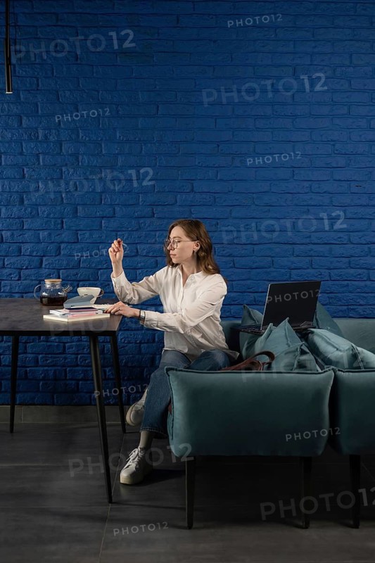 A woman wearing casual clothes works remotely in a cozy cafe. With a laptop, books, and tea, she is focused and productive, surrounded by a stylish blue brick wall