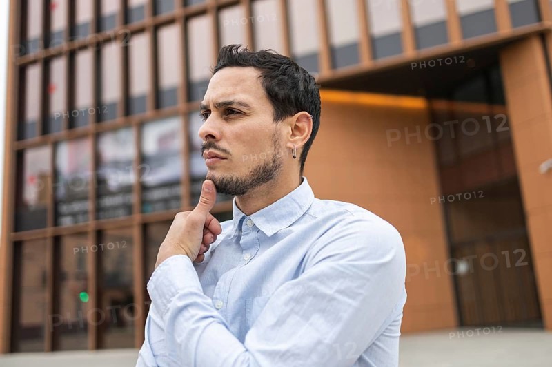A young man in a light blue shirt stands in front of a modern glass building, touching his chin in a thoughtful pose. His serious expression conveys deep thinking, confidence, and contemplation