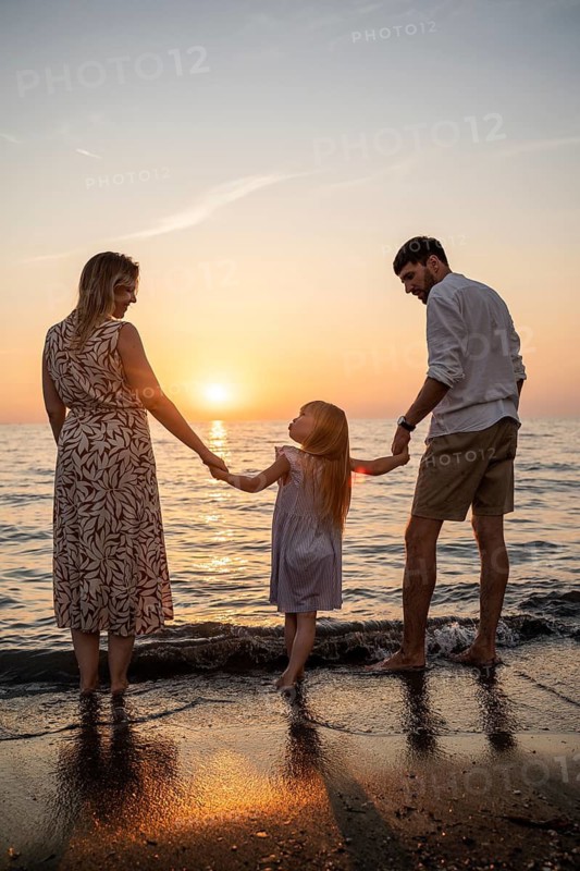 A loving family stands at the edge of the water, holding hands and watching the sun set over the ocean in Italy. A serene and heartfelt moment of connection and togetherness