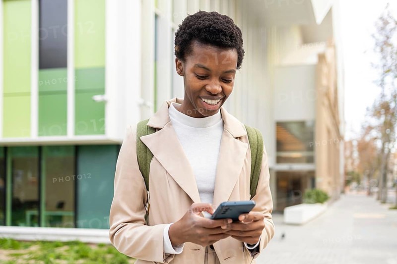 Smiling black woman using her smartphone in an urban setting, showcasing modern lifestyle and connectivity She stands in front of contemporary architecture, exuding confidence