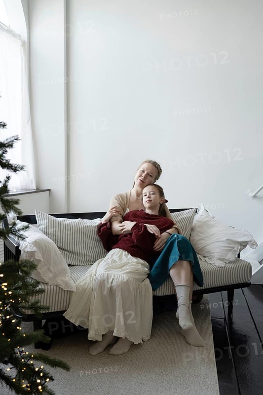 A mother and daughter share a warm embrace on a cozy striped sofa, with a softly lit Christmas tree nearby The serene setting captures the spirit of family and holiday joy