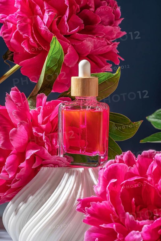 A glass dropper bottle filled with vibrant pink serum is surrounded by lush, blooming peonies. The backdrop is a deep blue, highlighting the elegant floral arrangement
