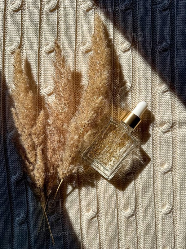 A glass serum cosmetic bottle with golden flakes sits on a textured cream knitted fabric beside pampas grass, illuminated by sunlight in the bathroom. Perfect for beauty and wellness themes