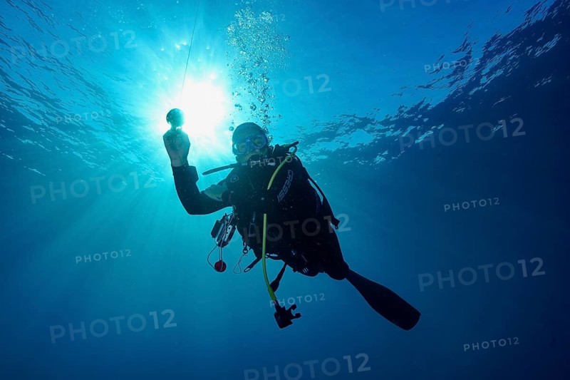 A female diver in full gear floats underwater while sunbeams shine through the water during a safety stop, Giens Peninsula, Mediterranean, Porquerolles, Provence, Côte d'Azur, France