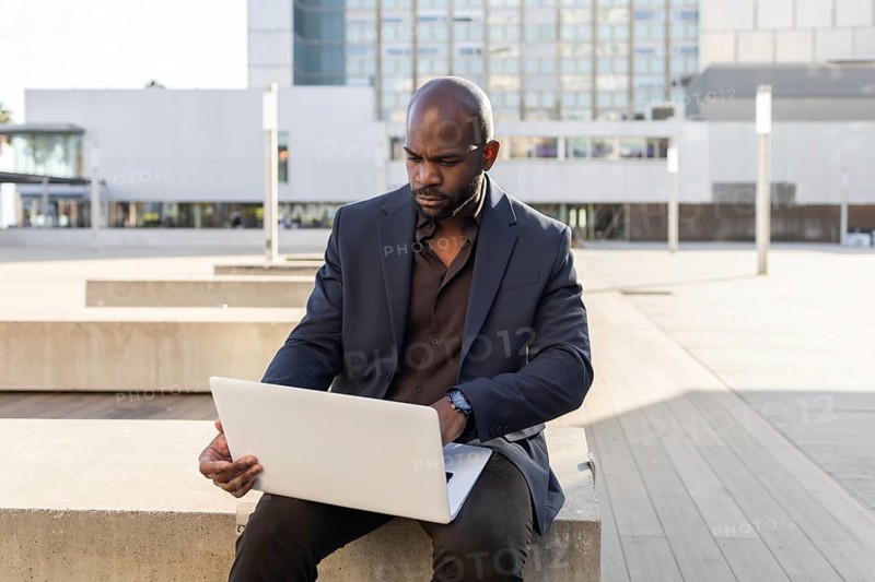 A business cuban man in a dark suit sits outdoors, focused on his laptop. The urban backdrop with a modern building adds a professional and dynamic feel to the scene