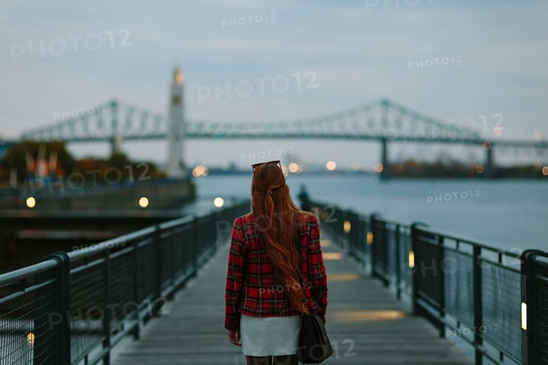 Back view of unrecognizable woman with red hair walking on a boardwalk in Montreal during fall, with a river view and iconic bridge in the background
