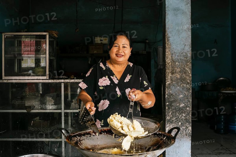 A woman smiles while cooking in a rustic kitchen, holding a frying pan filled with a traditional dish. Natural light highlights her joyful expression and vibrant attire