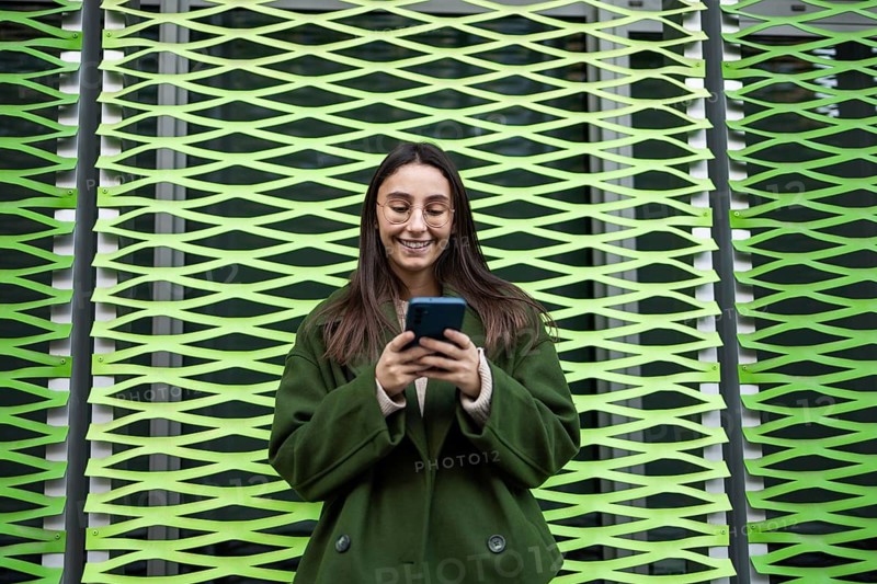 A woman wearing a green coat happily uses her smartphone while standing in front of a vibrant green patterned wall The scene captures a modern urban lifestyle