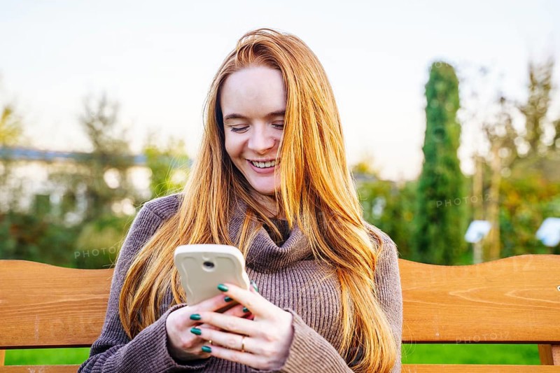 Beautiful red-haired pregnant woman sitting on a park bench, smiling as she looks at her smartphone. The serene outdoor setting enhances the sense of tranquility and joy