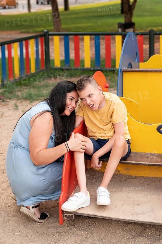 Mother and son with cerebral palsy share a tender moment at a colorful playground. Smiling and bonding, they reflect love, care, and warmth in an inclusive outdoor setting