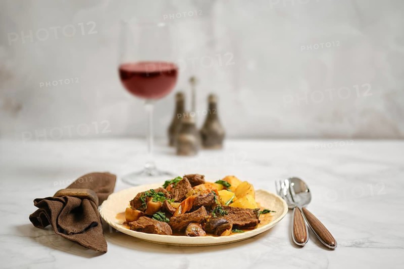 A plate holds pieces of cooked beef and roasted potatoes. A glass of red wine sits nearby. The setting includes a napkin and utensils. The overall look is simple and inviting
