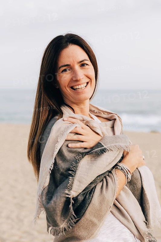 A cheerful woman wrapped in a cozy scarf standing on the beach, smiling sincerely while enjoying the seaside ambiance looking at camera