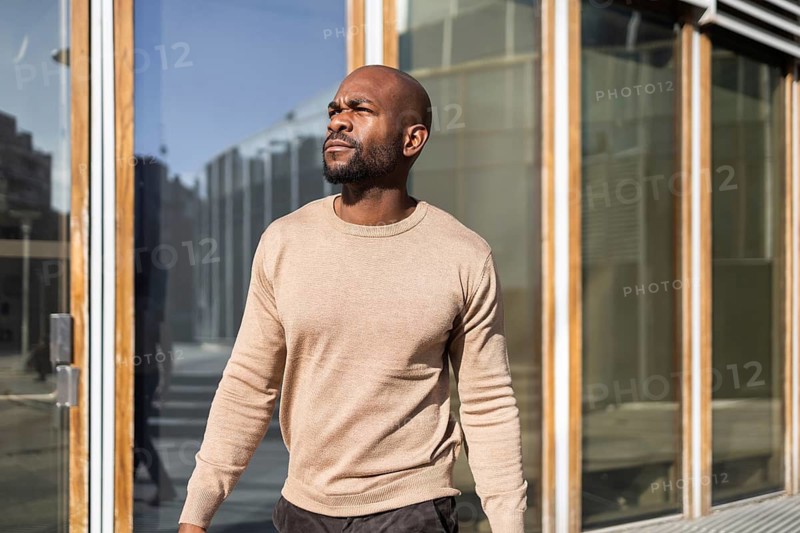 A cuban man strides confidently outside, basking in sunlight next to a modern glass structure. His casual beige sweater adds to the relaxed urban ambiance of the scene