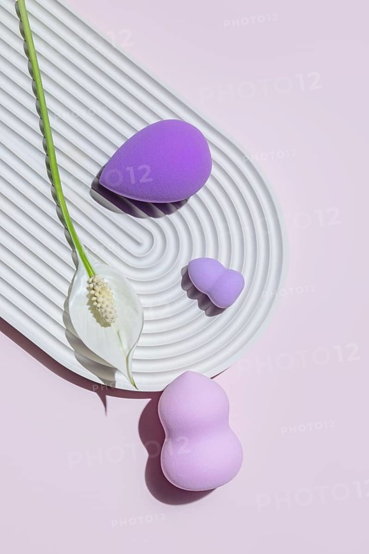 Top view of assorted makeup egg sponges, including beauty blenders, displayed creatively on a sculpted white surface with a delicate white flower, against a soft pink background