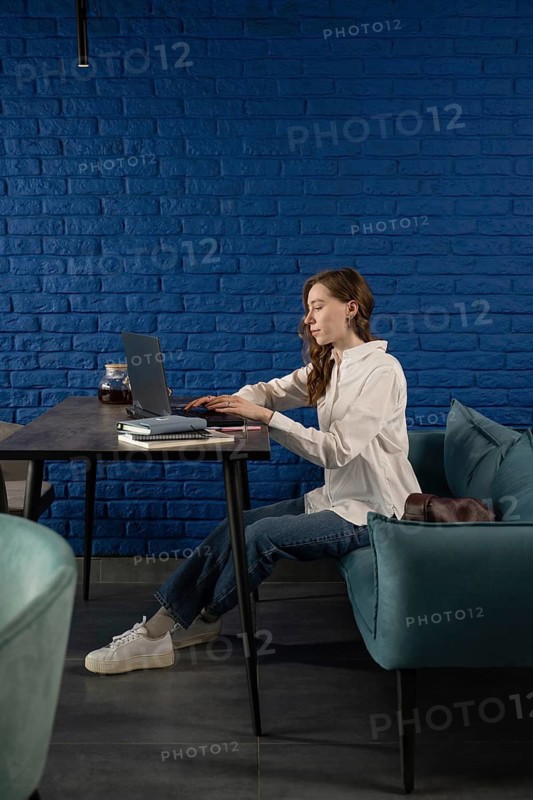 A woman sits in a modern blue cafe, working on a laptop at a black table. She is focused, wearing a white shirt and jeans, with notebooks and tea nearby