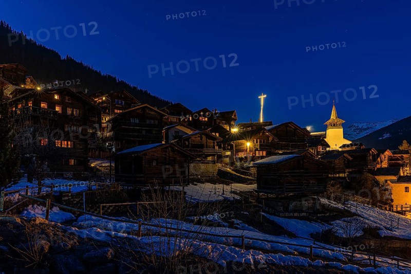 Snow-covered roofs of old farmhouses and barn storage, in the back the illuminated cross and the illuminated church tower of the Saint Theodule church, night view, historic town center, Grimentz, Val d'Anniviers, Valais Alps, Canton of Valais, Switzerland