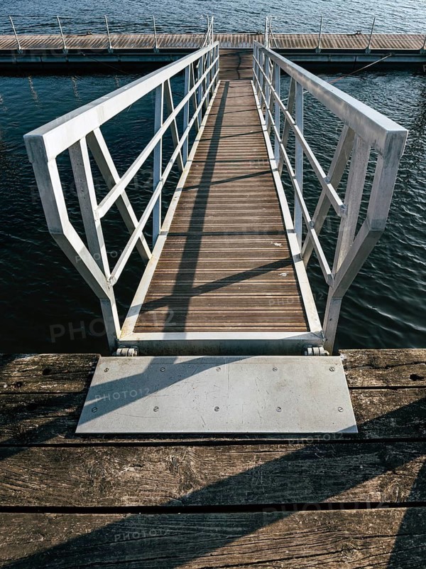A wooden dock pathway, enclosed by a metal railing, leads to a floating pier over calm water. The perspective captures the shadows and reflections, emphasizing the peaceful and serene setting
