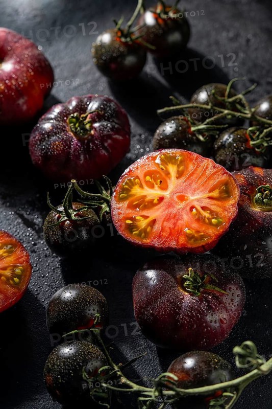 Close up of colorful heirloom tomatoes, both whole and halved, with water droplets. The rich textures and vibrant colors contrast beautifully against the dark backdrop
