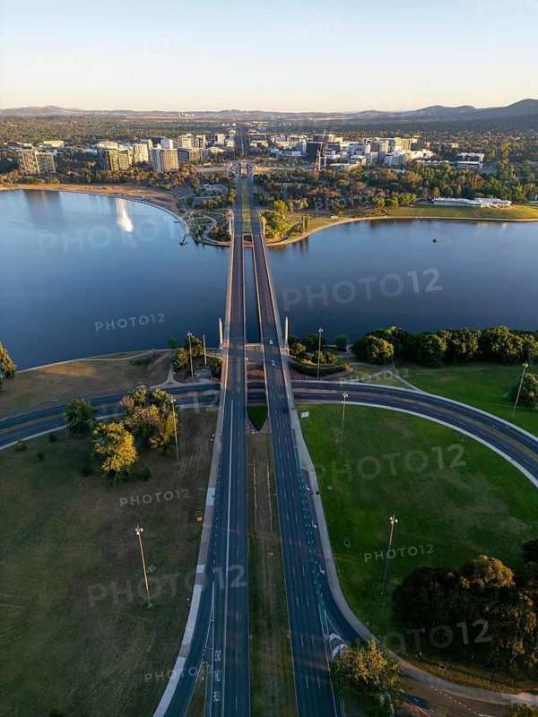 From above, a drone captures the stunning cityscape of Canberra, Australia, with a prominent highway bridge crossing the large Molonglo River, surrounded by lush greenery and the urban skyline at dawn